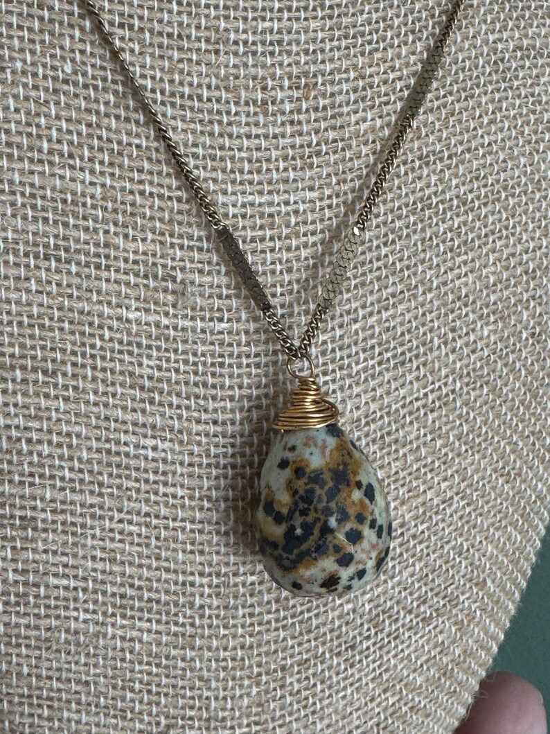 Puede incluir: Un collar con colgante en forma de l&aacute;grima. El colgante es una piedra moteada, envuelta con alambre dorado. La cadena es de color dorado. El collar se exhibe sobre una superficie texturizada de color neutro.