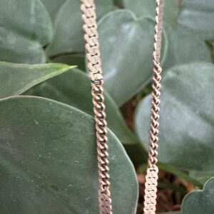 Puede incluir: Primer plano de dos collares dorados con diferentes estilos de cadena. Uno presenta un dise&ntilde;o de eslabones planos y rectangulares, mientras que el otro tiene una cadena de cuerda trenzada cl&aacute;sica. Los collares est&aacute;n sobre un fondo de hojas verdes.