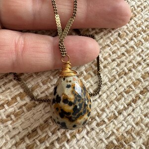 Puede incluir: Un collar con colgante en forma de l&aacute;grima. El colgante es una piedra moteada con manchas negras y marrones, envuelta en alambre dorado. La cadena es de color dorado delicado.