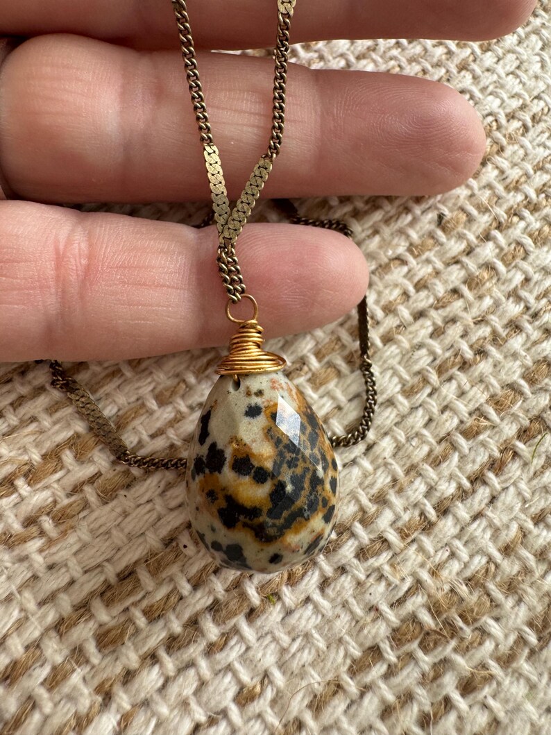 Puede incluir: Un collar con colgante en forma de l&aacute;grima. El colgante es una piedra moteada con manchas negras y marrones, envuelta en alambre dorado. La cadena es de color dorado delicado.