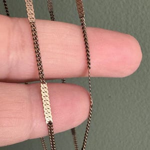 Puede incluir: Primer plano de un delicado collar dorado. El collar presenta una combinaci&oacute;n de peque&ntilde;as cadenas estrechamente unidas y segmentos planos y rectangulares. El dise&ntilde;o es simple y elegante, con una est&eacute;tica vintage.