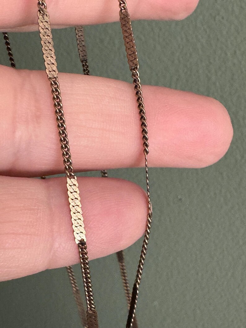 Puede incluir: Primer plano de un delicado collar dorado. El collar presenta una combinaci&oacute;n de peque&ntilde;as cadenas estrechamente unidas y segmentos planos y rectangulares. El dise&ntilde;o es simple y elegante, con una est&eacute;tica vintage.