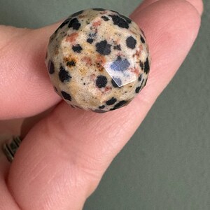 Puede incluir: Una gema esf&eacute;rica facetada con un patr&oacute;n moteado de puntos negros sobre un fondo beige y marr&oacute;n rojizo. La piedra se sostiene entre dos dedos, mostrando su superficie pulida y su coloraci&oacute;n &uacute;nica.