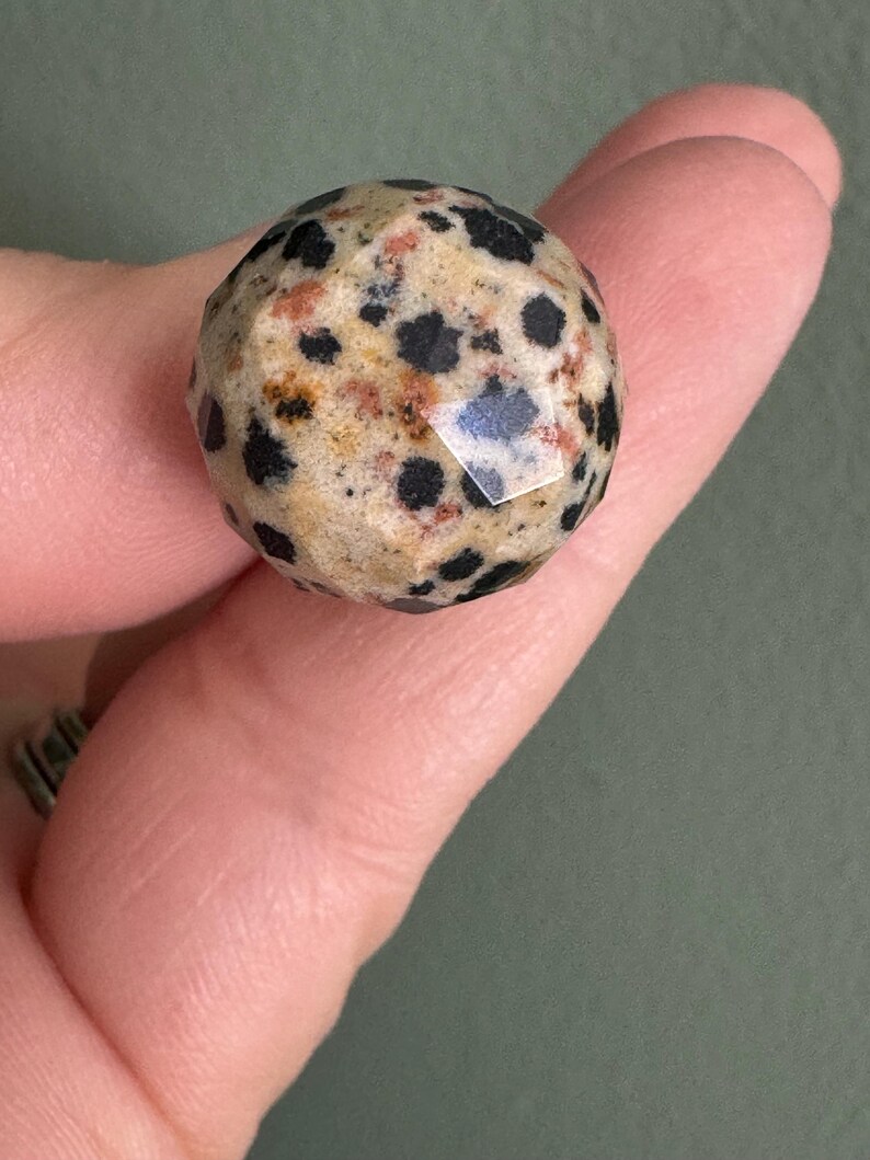 Puede incluir: Una gema esf&eacute;rica facetada con un patr&oacute;n moteado de puntos negros sobre un fondo beige y marr&oacute;n rojizo. La piedra se sostiene entre dos dedos, mostrando su superficie pulida y su coloraci&oacute;n &uacute;nica.