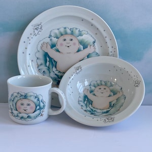 May include: A white ceramic children's dish set featuring a Cabbage Patch Kid doll design. The set includes a bowl, a plate, and a mug. Each piece features a blue and green cabbage leaf design with a Cabbage Patch Kid doll inside.