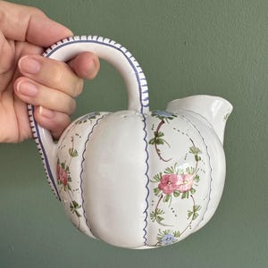 May include: White ceramic pitcher with a rounded body and a curved handle. The pitcher is decorated with pink and blue floral patterns and blue scalloped edges. The handle has blue stripes. The pitcher has a spout for pouring.