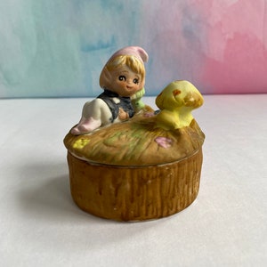 May include: A ceramic trinket box with a whimsical design featuring a small elf with blonde hair and a pink hat, sitting on a mushroom with a yellow dog beside them. The box is decorated with a brown tree trunk design.