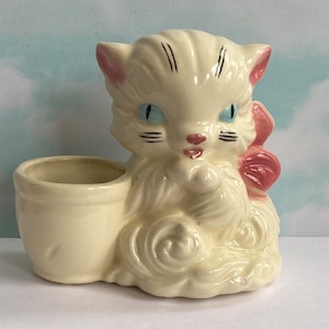 May include: A white ceramic planter shaped like a cat with a pink bow. The planter has blue eyes and black whiskers.