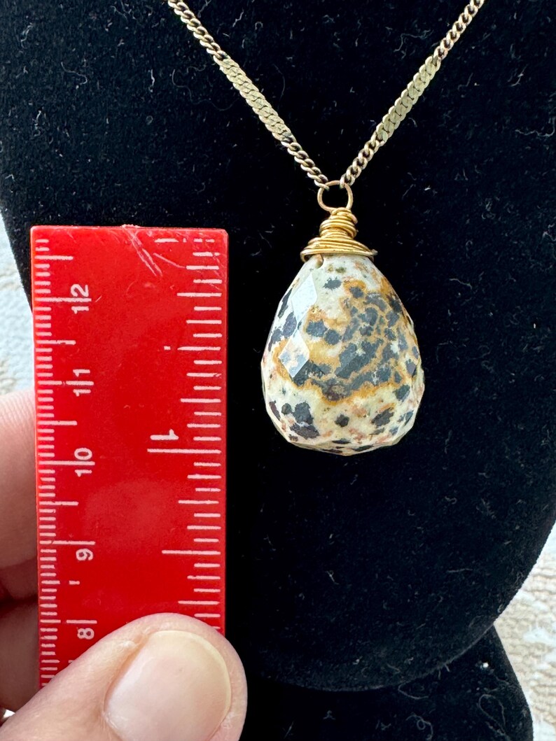 Puede incluir: Collar con colgante en forma de l&aacute;grima y cadena dorada. El colgante, de unos 3,2 cm de largo, es una piedra moteada, envuelta en alambre dorado. El collar se exhibe sobre un fondo de terciopelo negro.