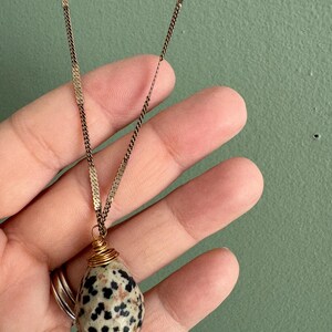 Puede incluir: Collar con colgante de piedra moteada. La piedra beige tiene manchas negras, est&aacute; envuelta en alambre dorado y cuelga de una delicada cadena dorada. El collar se muestra sobre un fondo verde.