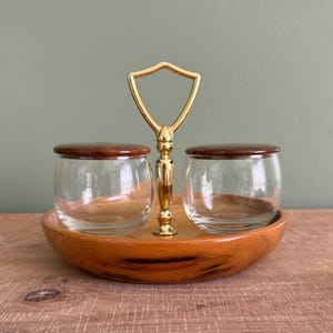 May include: A vintage condiment set featuring two clear glass jars with wooden lids, set on a wooden tray. A gold-colored handle connects the tray to the jars. The set is designed for serving condiments or small portions.