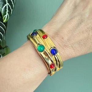 May include: A gold-tone metal cuff bracelet featuring a wavy, organic surface texture. The bracelet is embellished with several smooth, round cabochon stones in vibrant primary colors, including red, blue, and green, set into the metal surface.