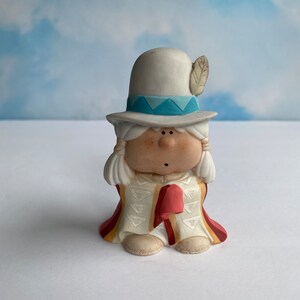 May include: A small ceramic figurine of a person wearing a white hat with a blue band and a feather, a white and red robe with geometric patterns, and white boots. The figurine has a round face with painted eyes, a nose, and a mouth.