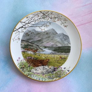 May include: A white ceramic plate with a gold rim features a painted scene of a mountain landscape with a lake, deer, and a grouse. The plate is decorated with a floral border.