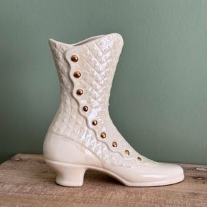 May include: A white ceramic boot-shaped vase with gold accents. The vase has a quilted design and is perfect for displaying flowers.