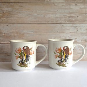 May include: Two off-white ceramic mugs with a speckled finish. Each mug features a detailed illustration of a bird with brown, yellow, and red plumage perched on a branch with orange flowers. The mugs have large, curved handles.