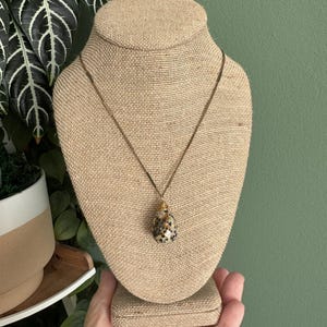 Puede incluir: Un collar con colgante de piedra en forma de l&aacute;grima, con un patr&oacute;n moteado en negro y beige. El colgante cuelga de una fina cadena dorada. El collar se exhibe en un expositor de joyer&iacute;a cubierto de arpillera.