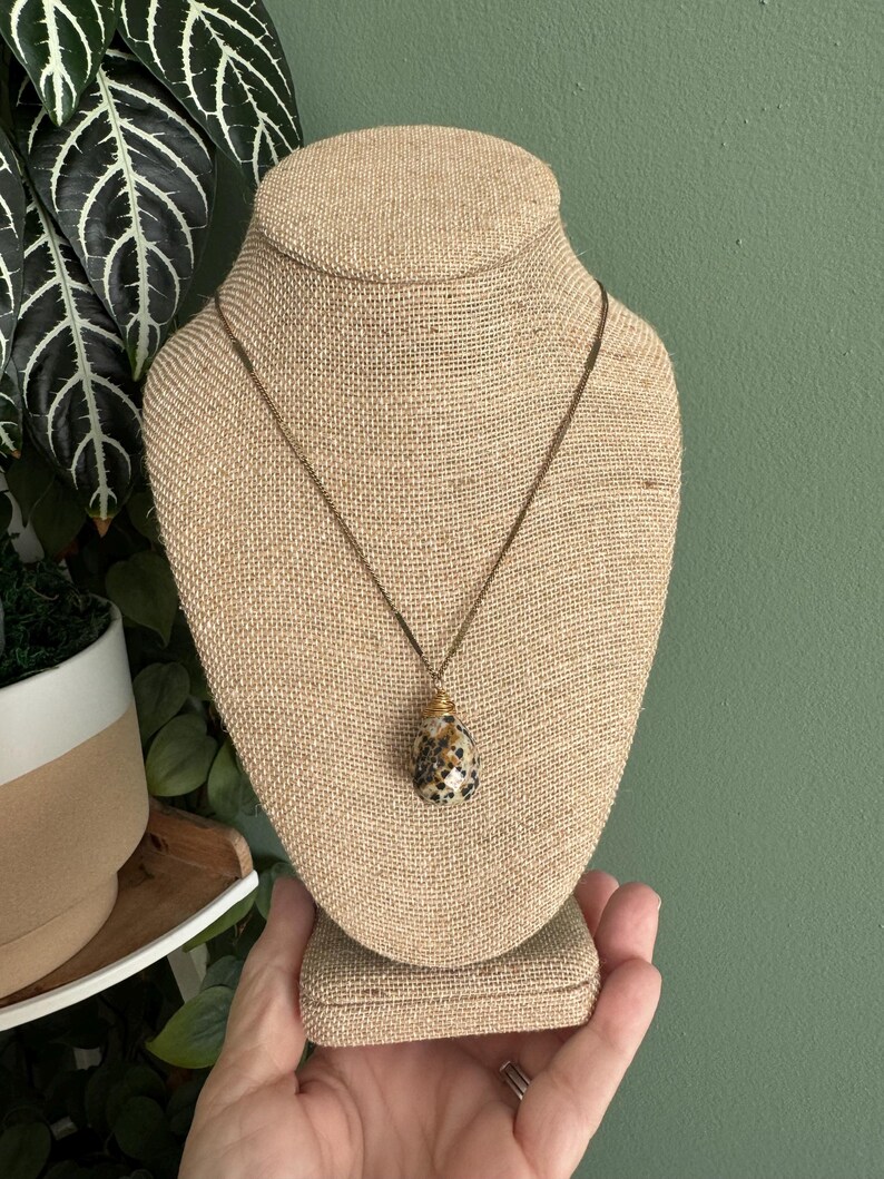 Puede incluir: Un collar con colgante de piedra en forma de l&aacute;grima, con un patr&oacute;n moteado en negro y beige. El colgante cuelga de una fina cadena dorada. El collar se exhibe en un expositor de joyer&iacute;a cubierto de arpillera.