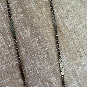 Puede incluir: Primer plano de dos collares dorados con un dise&ntilde;o de cadena rectangular texturizada. Los collares se muestran sobre una superficie de madera marr&oacute;n claro. Las cadenas tienen una apariencia delicada y vintage.