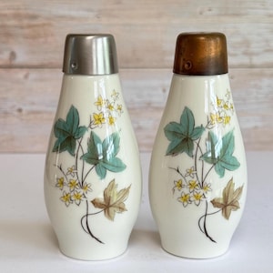 May include: A pair of white ceramic salt and pepper shakers with a tapered shape. Each shaker features a botanical illustration of green leaves and small yellow flowers. One shaker has a silver-toned metal cap, while the other has a copper-toned cap.