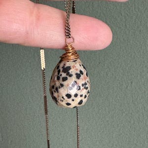 Puede incluir: Collar con colgante de jaspe d&aacute;lmata. La piedra en forma de l&aacute;grima presenta un patr&oacute;n moteado de manchas negras sobre un fondo crema. La piedra est&aacute; envuelta en alambre de color dorado y cuelga de una delicada cadena.