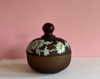 May include: A brown, round jar with a matching lid and a small, round handle. The jar is decorated with white daisies and green leaves. The jar is sitting on a white surface with a pink background.