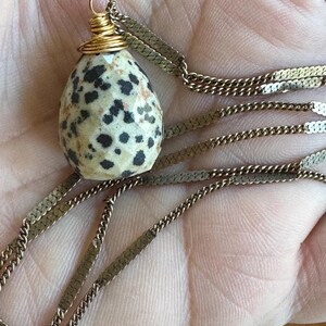 Puede incluir: Un collar con un colgante de piedra moteada en forma de huevo con manchas negras, envuelto en alambre de oro. El colgante cuelga de una delicada cadena plana de oro. Una pieza de joyer&iacute;a &uacute;nica.