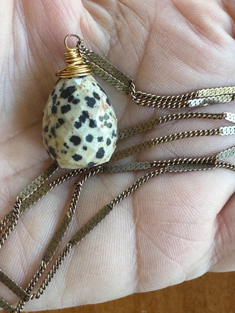 Puede incluir: Un collar con un colgante de piedra moteada en forma de huevo con manchas negras, envuelto en alambre de oro. El colgante cuelga de una delicada cadena plana de oro. Una pieza de joyer&iacute;a &uacute;nica.