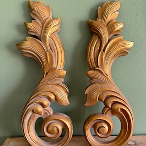 May include: Two ornate, gold-colored wall decor pieces. Each piece features a scrolling design with detailed leaf and swirl patterns. The pieces are symmetrical and have a vintage aesthetic, perfect for adding a touch of elegance to any space.