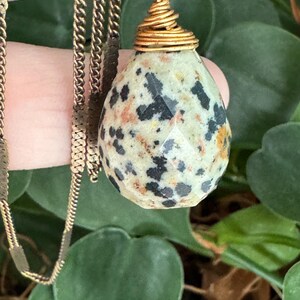 Puede incluir: Un collar de cadena dorada con un colgante en forma de l&aacute;grima. El colgante es una piedra beige claro con motas negras y naranjas, facetada para captar la luz. El colgante est&aacute; envuelto en alambre dorado.