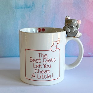 May include: White ceramic mug with a gray hippopotamus figurine holding a lollipop on the handle. The mug has red text that reads "The Best Diets Let You Cheat A Little!"