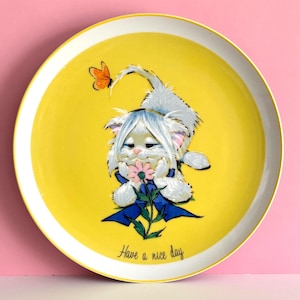 May include: A round yellow ceramic plate featuring a white fluffy kitten with blue eyes holding a pink flower. A small orange butterfly is positioned above the kitten's head. The plate has a blue ribbon graphic and the text Have a nice day.