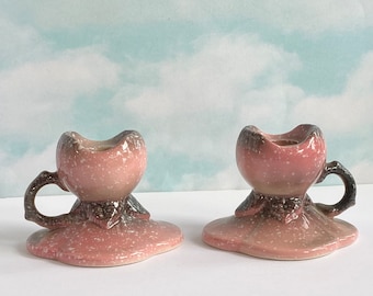 May include: Two pink and brown ceramic candle holders with a speckled glaze. Each holder has a small cup for a candle and a handle.