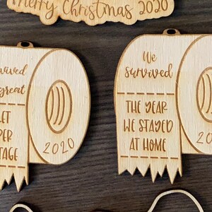 May include: Two wooden ornaments shaped like toilet paper rolls. The first ornament says "I survived the great toilet paper shortage 2020". The second ornament says "We survived the year we stayed at home 2020".