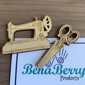 Puede incluir: Dos suministros de artesanía de madera, una máquina de coser y un par de tijeras, ambos con detalles intrincados. La máquina de coser tiene un diseño floral y las tijeras tienen un diseño clásico.