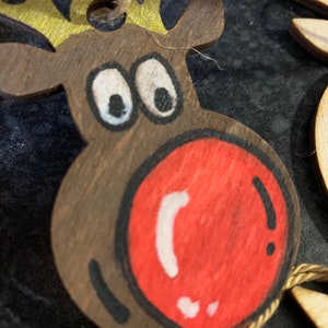 May include: A wooden reindeer ornament with a red nose, black eyes, and gold antlers. The ornament is painted with white details and has a string for hanging.