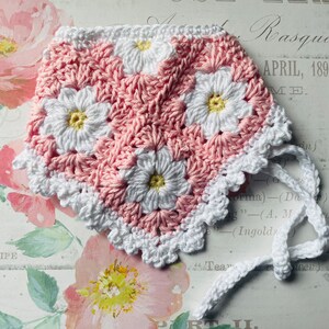 Op de afbeelding: Een roze en witte gehaakte baby muts met een witte gehaakte strik. De muts heeft een madeliefjespatroon.
