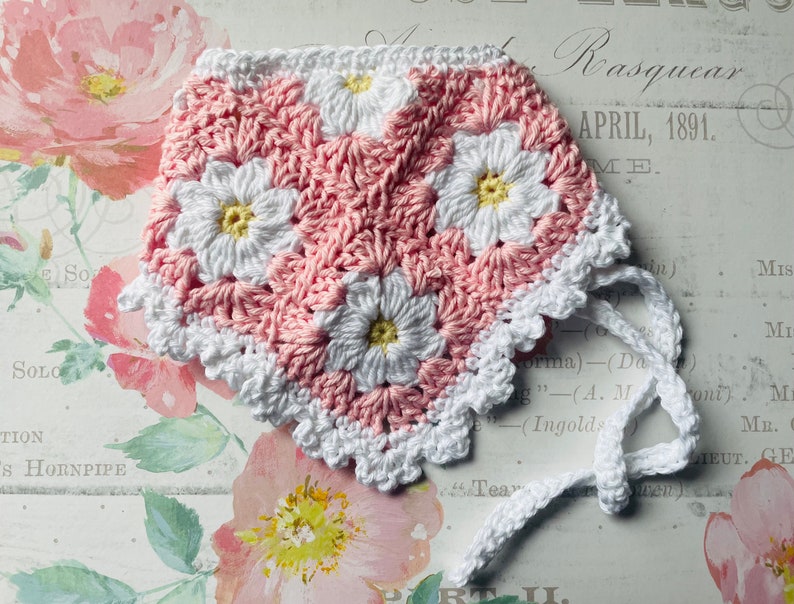Op de afbeelding: Een roze en witte gehaakte baby muts met een witte gehaakte strik. De muts heeft een madeliefjespatroon.