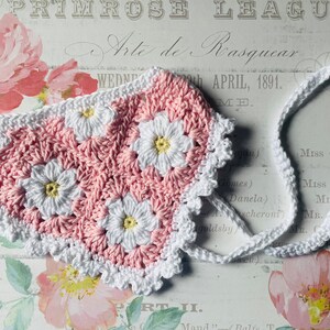 Op de afbeelding: Een roze en witte gehaakte baby muts met een witte gehaakte rand. De muts heeft een madeliefjespatroon.