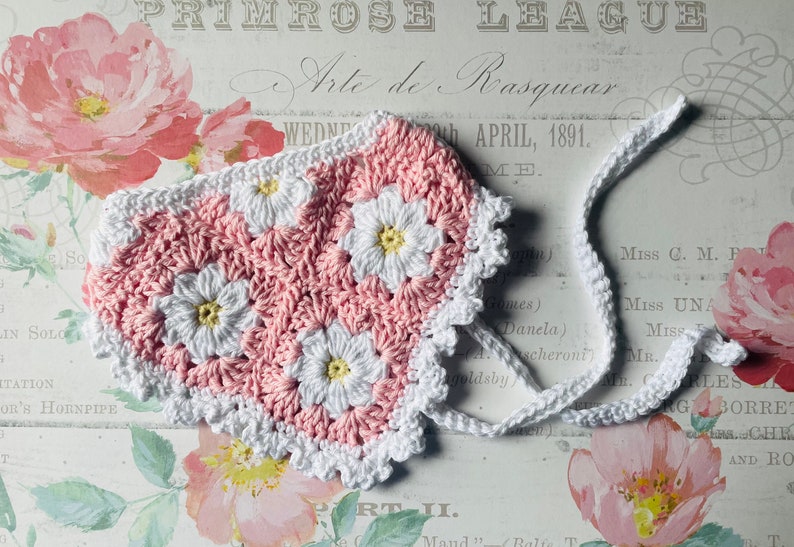 Op de afbeelding: Een roze en witte gehaakte baby muts met een witte gehaakte rand. De muts heeft een madeliefjespatroon.