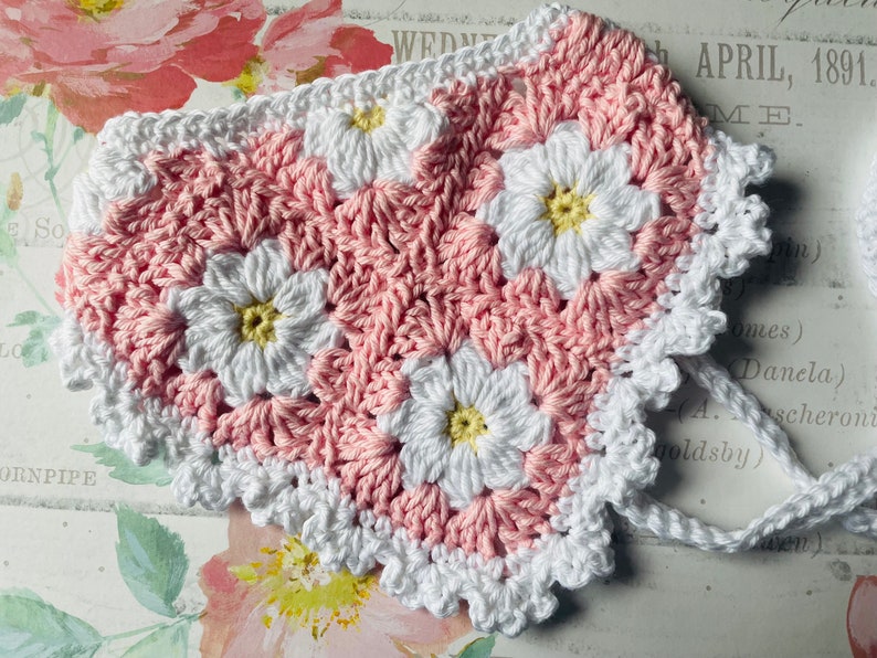 Op de afbeelding: Een roze en wit geh&auml;keld hartvormig stuk met een witte geh&auml;kelde rand. Het stuk is gemaakt van kleine bloemen met gele centra.