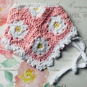 Op de afbeelding: Een roze en witte gehaakte baby muts met een witte gehaakte strik. De muts heeft een madeliefjespatroon.