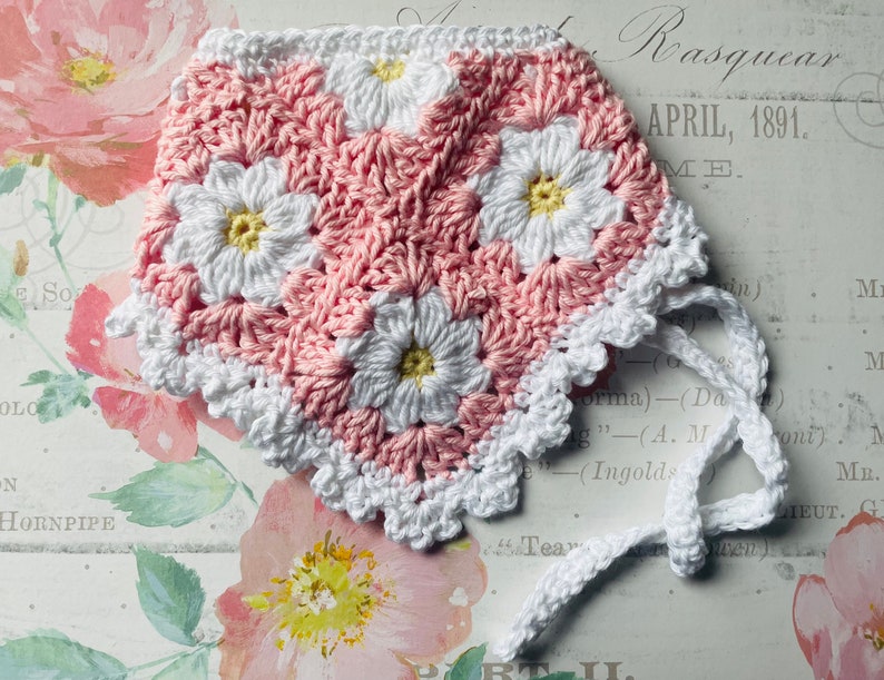 Op de afbeelding: Een roze en witte gehaakte baby muts met een witte gehaakte strik. De muts heeft een madeliefjespatroon.
