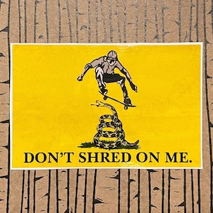 K&ouml;nnte beinhalten: Gelber Aufkleber mit einem Skateboarder, der &uuml;ber einer zusammengerollten Schlange springt. Der Text "DON'T SHRED ON ME" ist in Schwarz gedruckt. Der Skateboarder und die Schlange sind in Schwarzwei&szlig;.