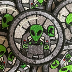 May include: Close-up of a green alien embroidered patch. The alien is wearing a black helmet and is sitting in a cockpit. The patch is circular and has a gray and white background.