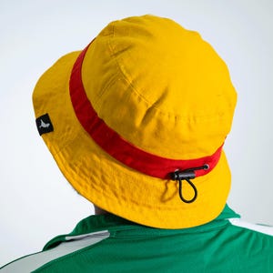 May include: A yellow bucket hat with a red band and a black adjustable strap. The hat has a small black logo on the side.