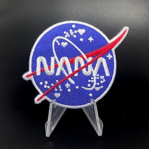 May include: A blue and red embroidered patch with the word "NASA" in white lettering. The patch has a white rocket ship design and a red and white swirl design.