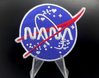 Könnte beinhalten: Ein blauer und roter gestickter Aufnäher mit dem Wort "NASA" in weißen Buchstaben. Der Aufnäher zeigt ein weißes Raketenschiffdesign und ein rotes und weißes Wirbeldesign.