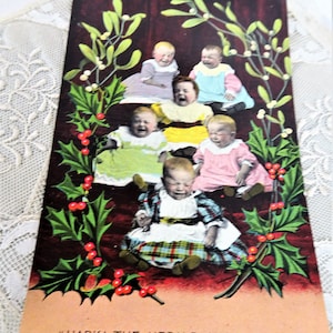 Peut inclure: Une carte postale vintage représentant six bébés vêtus de différents costumes, assis en cercle sur un fond de houx vert et de baies rouges. Le texte "HARK! THE HERALD ANGELS SING" est imprimé en bas de la carte postale.