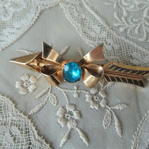 May include: A gold-toned arrow brooch with a blue rhinestone bow. The arrow is detailed with a textured design and the bow is tied with a knot.